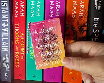 May include: A miniature book keychain titled A Court of Splintered Harmony by Sarah J. Maas, featuring a gradient orange and pink cover with star illustrations, held in front of a row of full-sized books with colorful spines by the same author.