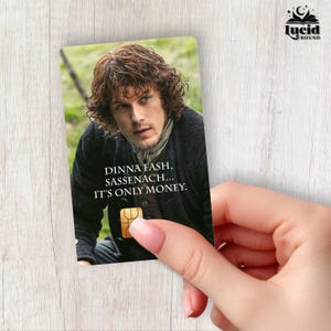 May include: A rectangular card featuring a portrait of a person with wavy brown hair wearing a dark jacket and a scarf. The text reads DINNA FASH, SASSENACH... IT'S ONLY MONEY. A gold-coloured rectangular chip is visible at the bottom centre.
