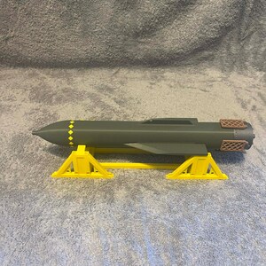 May include: A gray model missile with yellow diamond accents and a brown textured tail, resting on a yellow stand. The missile is a cylindrical shape with a pointed nose.