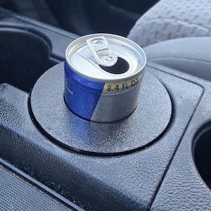 May include: An open Red Bull energy drink can, 8.4 fl oz, sits in a car's cup holder. The can is silver with blue and red branding. The cup holder is black and textured, with a circular design to fit the can.