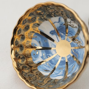 May include: A scalloped ceramic bowl featuring a gold-rimmed edge and a gold-colored center circle. The interior displays alternating panels of a blue and white marbled pattern and a textured, reflective gold finish with a wavy, embossed surface.