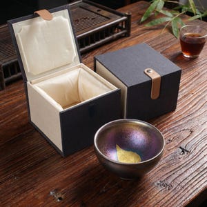 May include: A metallic tea bowl with a gold leaf design, presented with a black gift box. The box has a beige interior and a leather strap closure. The bowl has iridescent colors and sits on a wooden surface.