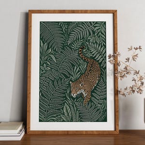 May include: A framed art print featuring a spotted jaguar walking through a dense forest of white fern leaves on a dark green background. The artwork is enclosed in a light brown wooden frame, showcasing a detailed illustration of the wild cat.