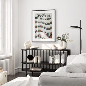 May include: A framed "The New Yorker" magazine cover art print featuring a colorful illustration of a museum interior. The print hangs above a black console table with decorative objects and books. A modern lamp and white sofa are also visible.