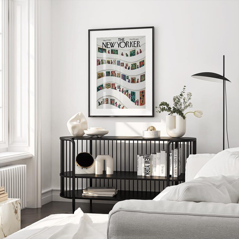 May include: A framed "The New Yorker" magazine cover art print featuring a colorful illustration of a museum interior. The print hangs above a black console table with decorative objects and books. A modern lamp and white sofa are also visible.
