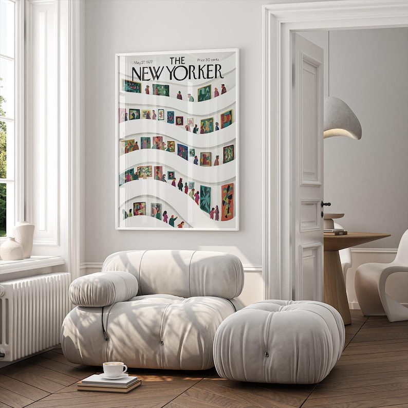 May include: A framed "The New Yorker" magazine cover from May 23, 1977, featuring an art gallery scene with colorful paintings and figures. The artwork is displayed in a bright, modern living room with a white sofa and ottoman.