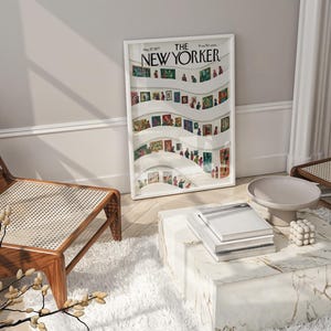 May include: A framed "The New Yorker" magazine cover art print featuring a colorful illustration of people in an art gallery. The print is displayed in a white frame, resting against a wall. A modern living room setting with a marble coffee table and wooden chairs.
