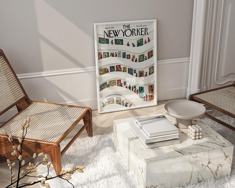 May include: A framed "The New Yorker" magazine cover art print featuring a colorful illustration of people in an art gallery. The print is displayed in a white frame, resting against a wall. A modern living room setting with a marble coffee table and wooden chairs.