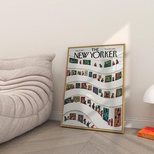 May include: A framed New Yorker magazine cover featuring an art gallery scene with people viewing paintings. The cover is in a gold frame and is leaning against a wall. A beige chair and a white lamp are also in the image.