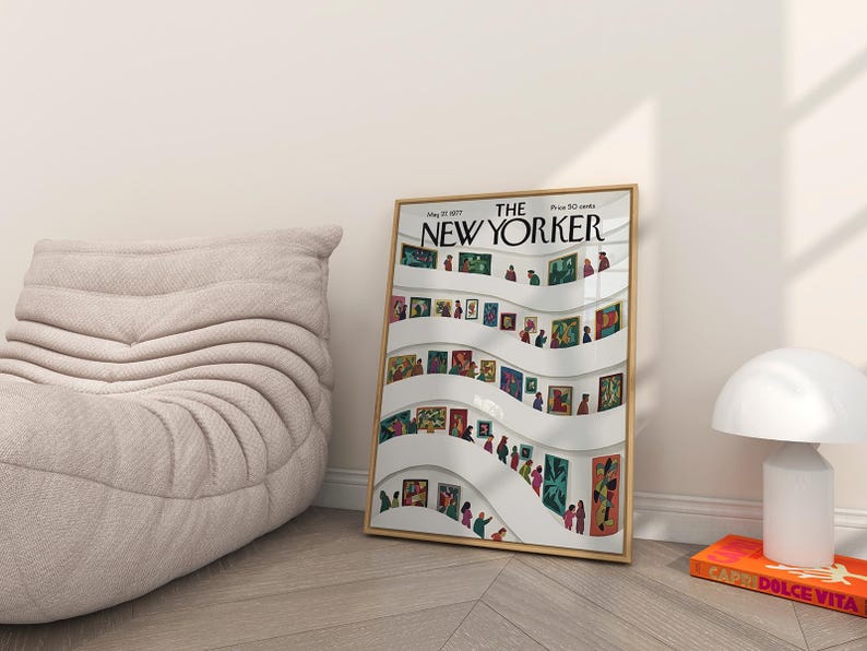 May include: A framed New Yorker magazine cover featuring an art gallery scene with people viewing paintings. The cover is in a gold frame and is leaning against a wall. A beige chair and a white lamp are also in the image.