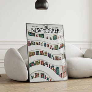 May include: A framed New Yorker magazine cover from May 27, 1977, featuring an art gallery scene with people viewing paintings. The artwork is displayed on white curved walls. The magazine is propped up against a white sofa on a wooden floor.