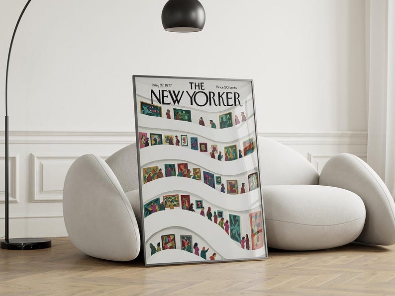May include: A framed New Yorker magazine cover from May 27, 1977, featuring an art gallery scene with people viewing paintings. The artwork is displayed on white curved walls. The magazine is propped up against a white sofa on a wooden floor.