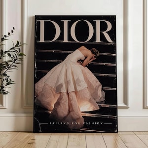 May include: A framed print with the word "DIOR" at the top and "FALLING FOR FASHION" at the bottom. It depicts a woman in a strapless gown on a staircase. The dress is a pale pink colour.