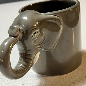 May include: Grey ceramic mug featuring an elephant head design. The elephant's trunk forms the handle. The mug has a glossy finish and a dark interior. The elephant's features are detailed, including eyes and wrinkles.