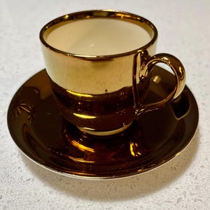 May include: A gold-coloured teacup and saucer set. The cup and saucer have a shiny, reflective surface. The interior of the cup is a light cream colour. The saucer has a starburst design on the edge.