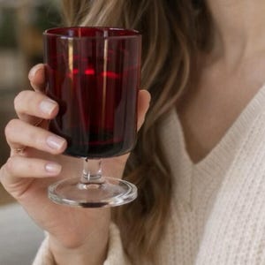 May include: A deep red wine glass with a clear stem and base. The glass is held by a person wearing a cream-coloured jumper. The glass is filled with a dark liquid, possibly wine or juice.