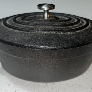 May include: A black cast iron Dutch oven with a lid and two side handles. The lid has a silver knob and concentric circular ridges. The pot is sitting on a white surface.