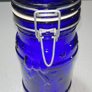 May include: A deep blue glass storage jar with a metal clasp and a white rubber seal. The jar has embossed floral patterns and a cylindrical shape. The lid is also blue. The jar is designed for storage.