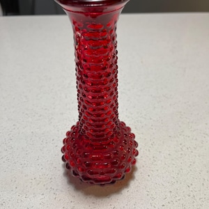 May include: A vibrant ruby red glass vase featuring a hobnail texture. The vase has a flared opening, a textured, slender neck, and a rounded base. The vase is approximately 18 cm tall, suitable for small floral arrangements or as a decorative item.