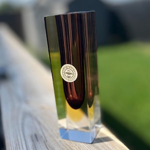 May include: A tall, rectangular glass vase featuring a deep purple, rounded interior cavity encased in clear, thick glass. A circular white sticker with the text Artigiano Muranese is affixed to the front surface of the glass object.