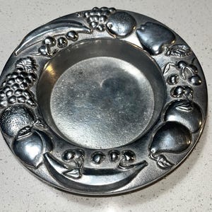 May include: A silver-coloured, round dish with a raised rim. The rim is decorated with embossed fruit, including grapes, apples, pears, bananas, and cherries. The dish has a shallow, circular centre.