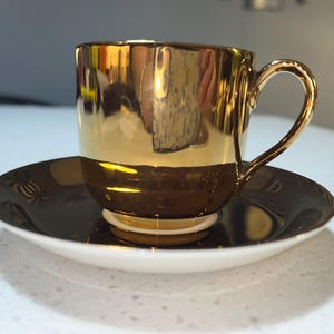 May include: A gold-coloured teacup and saucer set. The cup and saucer are two-toned, with a gold upper half and a dark brown lower half. The saucer is white on the bottom. The cup has a curved handle.