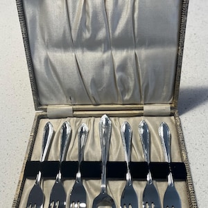 Könnte beinhalten: Ein Satz Silberbesteck in einem Vintage-Etui. Die Gabeln haben ein dekoratives Design an den Griffen und sind in einer cremefarbenen, satin gefütterten Box mit gemustertem Äußeren angeordnet. Das Etui ist geöffnet und zeigt das Besteck.