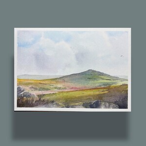 Könnte beinhalten: Aquarell-Landschaftsmalerei eines grünen und gelben Hügels unter einem bewölkten blauen Himmel. Das Kunstwerk zeigt einen Berg in der Ferne und einen Vordergrund aus Felsen und Vegetation. Das Gemälde ist mit einem weißen Rand gerahmt.