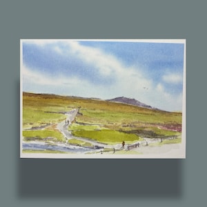 Könnte beinhalten: Aquarellmalerei einer Landschaft mit einem gewundenen Pfad, grünen Feldern und einem blauen Himmel mit Wolken. Ein kleiner Bach und ein Holzzaun sind zu sehen. Das Kunstwerk befindet sich auf einer weißen rechteckigen Karte.