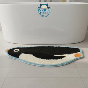 Könnte beinhalten: Eine pinguinförmige Badematte mit schwarzem Körper, weißem Bauch und orangefarbenen Akzenten. Die Matte hat einen hellblauen Rand und ist vor einer weißen Badewanne platziert. Auf der Matte steht der Text "Pro Rug Original".