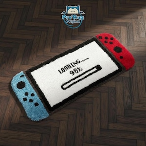 Peut inclure: Tapis rectangulaire en forme de manette de console de jeu. Il présente un écran blanc avec le mot « LOADING » et une barre de progression à 98 %. Les côtés de la manette sont bleus et rouges, avec des détails noirs. Le tapis est posé sur un sol en bois.
