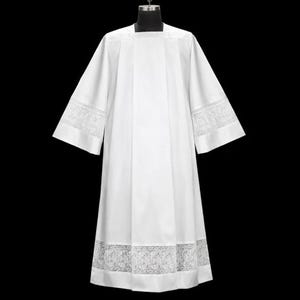 May include: A white liturgical alb with a square neckline and wide sleeves. The sleeves and hem feature delicate lace trim. The garment is displayed on a black mannequin, showcasing its full length and design.