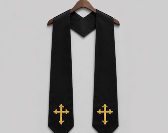 Puede incluir: Una estola litúrgica negra con cuello en V y dos paneles largos y rectos. Cada panel está bordado con un diseño de cruz de color dorado cerca de la parte inferior. La prenda se muestra en una percha de madera sobre un fondo neutro.