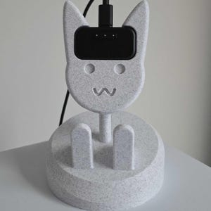 May include: A speckled grey cat-shaped charging station with a black charging port. The station has a round base with two vertical supports and a cat face design. The cat face has two round eyes and a carved-out smile. A black charging cable is plugged into the top.