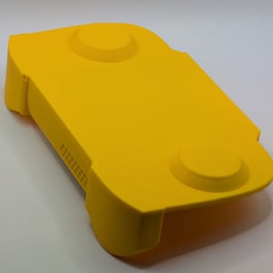 May include: A bright yellow handheld gaming console case with two circular raised buttons and a series of small rectangular vents. The case has rounded edges and a smooth, matte finish. The case is designed to protect the console.