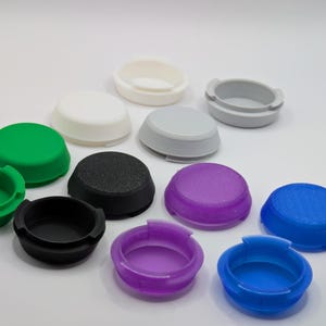 Peut inclure: Une collection de bouchons en plastique ronds et colorés de différentes nuances, dont le vert, le blanc, le gris, le noir, le violet et le bleu. Certains bouchons ont un dessus plat, tandis que d'autres ont un centre en creux. Les bouchons sont disposés sur une surface blanche.