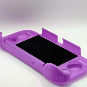 May include: A lavender-colored protective handheld gaming console case featuring a rectangular cutout for the screen and circular cutouts for buttons and joysticks, with a black felt-like lining on the interior base of the device holder.