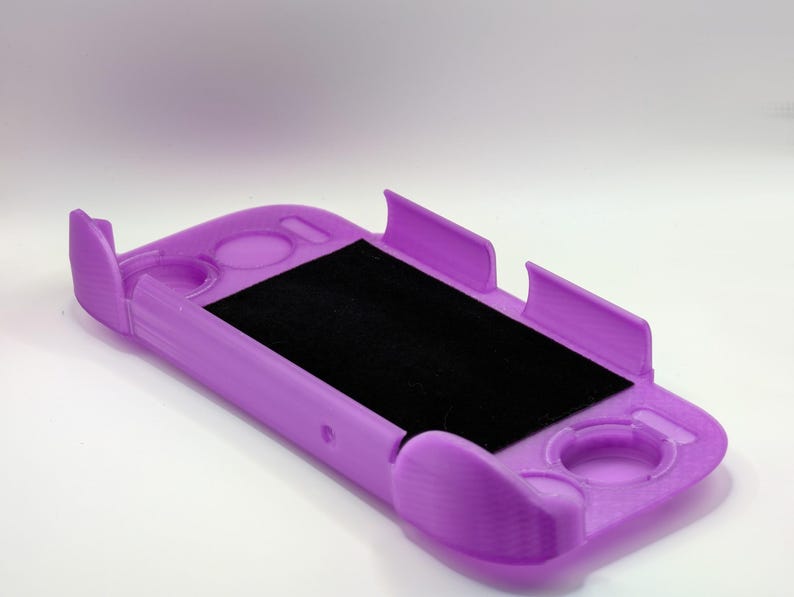 May include: A lavender-colored protective handheld gaming console case featuring a rectangular cutout for the screen and circular cutouts for buttons and joysticks, with a black felt-like lining on the interior base of the device holder.