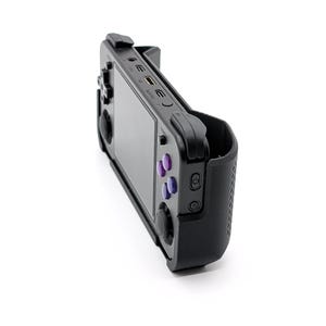 Puede incluir: Una consola de juegos portátil negra con pantalla y botones morados. El dispositivo está en una funda protectora negra y tiene varios puertos y botones visibles en los laterales. El dispositivo está ligeramente inclinado, mostrando su perfil lateral sobre un fondo blanco.