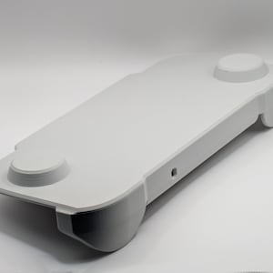 May include: A white, rectangular, handheld gaming controller grip featuring a smooth, matte surface, two circular raised button areas on the top panel, and ergonomic contoured handles on each side with a small circular port on the front edge.