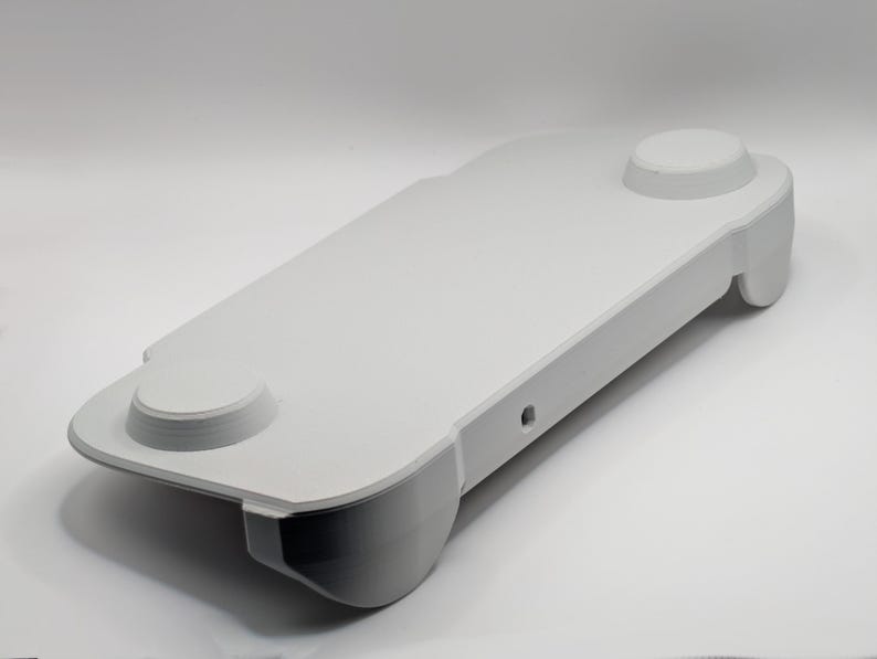 May include: A white, rectangular, handheld gaming controller grip featuring a smooth, matte surface, two circular raised button areas on the top panel, and ergonomic contoured handles on each side with a small circular port on the front edge.