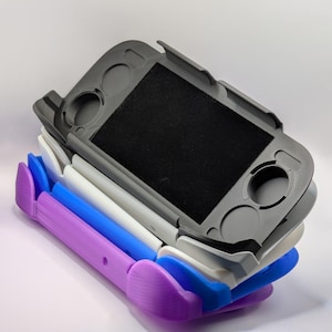 May include: A stack of four handheld gaming console protective cases in purple, blue, white, and dark gray. Each case features a rectangular center cutout, circular openings for analog sticks, and additional button slots, designed to fit a portable device.