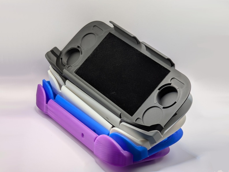 May include: A stack of four handheld gaming console protective cases in purple, blue, white, and dark gray. Each case features a rectangular center cutout, circular openings for analog sticks, and additional button slots, designed to fit a portable device.