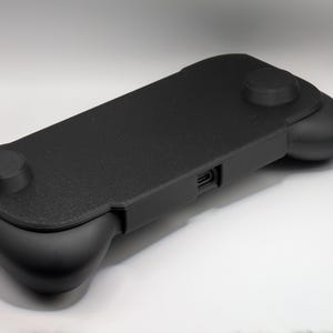 May include: A black handheld gaming controller grip featuring a textured rectangular center panel, two rounded side grips, and a central charging port opening. The device has a matte finish and a streamlined design for ergonomic handheld gaming.