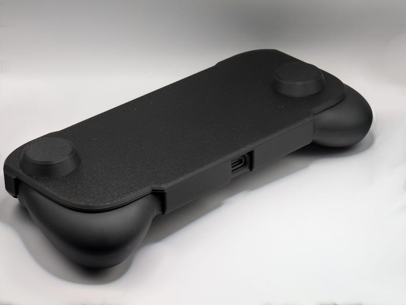 May include: A black handheld gaming controller grip featuring a textured rectangular center panel, two rounded side grips, and a central charging port opening. The device has a matte finish and a streamlined design for ergonomic handheld gaming.