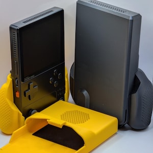 May include: A black handheld gaming console with a screen and directional pad, resting in a bright yellow ergonomic grip. Beside it stands a second black console in a dark grey grip, with a separate yellow protective cover placed in the foreground.