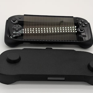 Pu&ograve; includere: Una console di gioco portatile nera con uno schermo che mostra una griglia di luci bianche e una base nera separata. Il dispositivo ha pulsanti di controllo e un D-pad. La base ha due impugnature circolari.