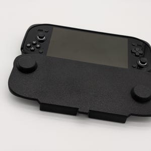 Pu&ograve; includere: Una console di gioco portatile nera con un'impugnatura nera strutturata. La console ha uno schermo e pulsanti di controllo. L'impugnatura ha due sporgenze circolari e una base rettangolare. Lo sfondo &egrave; bianco.