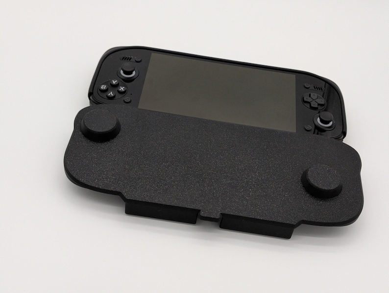 Pu&ograve; includere: Una console di gioco portatile nera con un'impugnatura nera strutturata. La console ha uno schermo e pulsanti di controllo. L'impugnatura ha due sporgenze circolari e una base rettangolare. Lo sfondo &egrave; bianco.