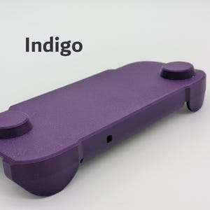 May include: A purple handheld gaming controller grip with a textured matte finish. The device features two circular thumbstick areas on the top surface and a small port on the side. The word Indigo is printed in dark gray text above the controller.